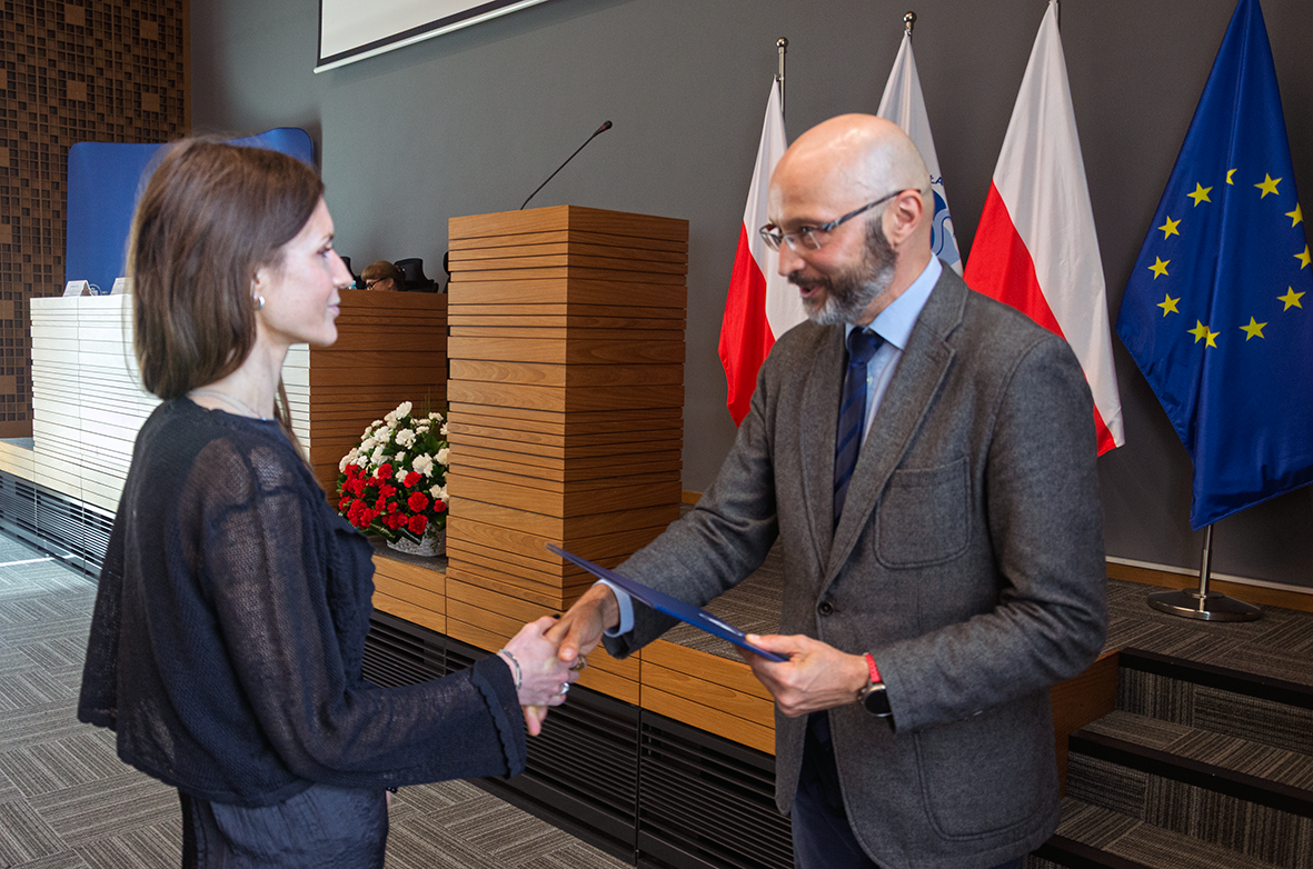 Na pierwszym planie widać dwie osoby — kobietę i mężczyznę — stojące naprzeciw siebie. Mężczyzna w garniturze i krawacie wręcza kobiecie dokument. Osoby podają sobie ręce w geście gratulacji. W tle stoją trzy flagi: dwie biało-czerwone (Polski) oraz jedna niebieska z żółtymi gwiazdami (Unia Europejska). Obok mównicy ustawiono bukiet biało-czerwonych kwiatów. Pomieszczenie jest przestronne, z nowoczesnym wystrojem i drewnianymi elementami dekoracyjnymi.