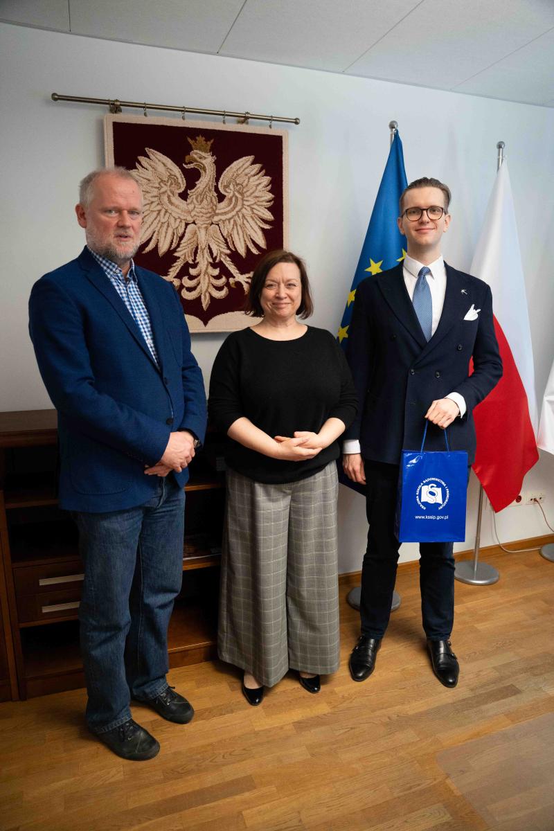Na zdjęciu widać trzy osoby stojące obok siebie w pomieszczeniu. Po lewej stronie stoi mężczyzna z brodą. Ma na sobie niebieską marynarkę, koszulę w kratę, dżinsy oraz czarne buty. Ręce trzyma złożone przed sobą. Na środku stoi kobieta z krótkimi, ciemnymi włosami. Ubrana jest w czarną bluzkę oraz szerokie, szare spodnie w kratę. Ma lekki uśmiech i trzyma dłonie złożone przed sobą. Po prawej stronie stoi młody mężczyzna w eleganckim, granatowym garniturze z krawatem. Ma okulary i zaczesane włosy w tył. W ręce trzyma niebieską torbę z białym logo i napisem Krajowej Szkoły Sądownictwa i Prokuratury. Na ścianie za nimi wisi czerwony materiał z wizerunkiem białego orła w koronie – symbolem Polski. Obok stoją dwie flagi na stojakach: flaga Unii Europejskiej (niebieska z żółtymi gwiazdami) oraz flaga Polski (biało-czerwona). Po lewej stronie widać drewnianą komodę. Podłoga jest drewniana, ściany jasne, a sufit z białych paneli.