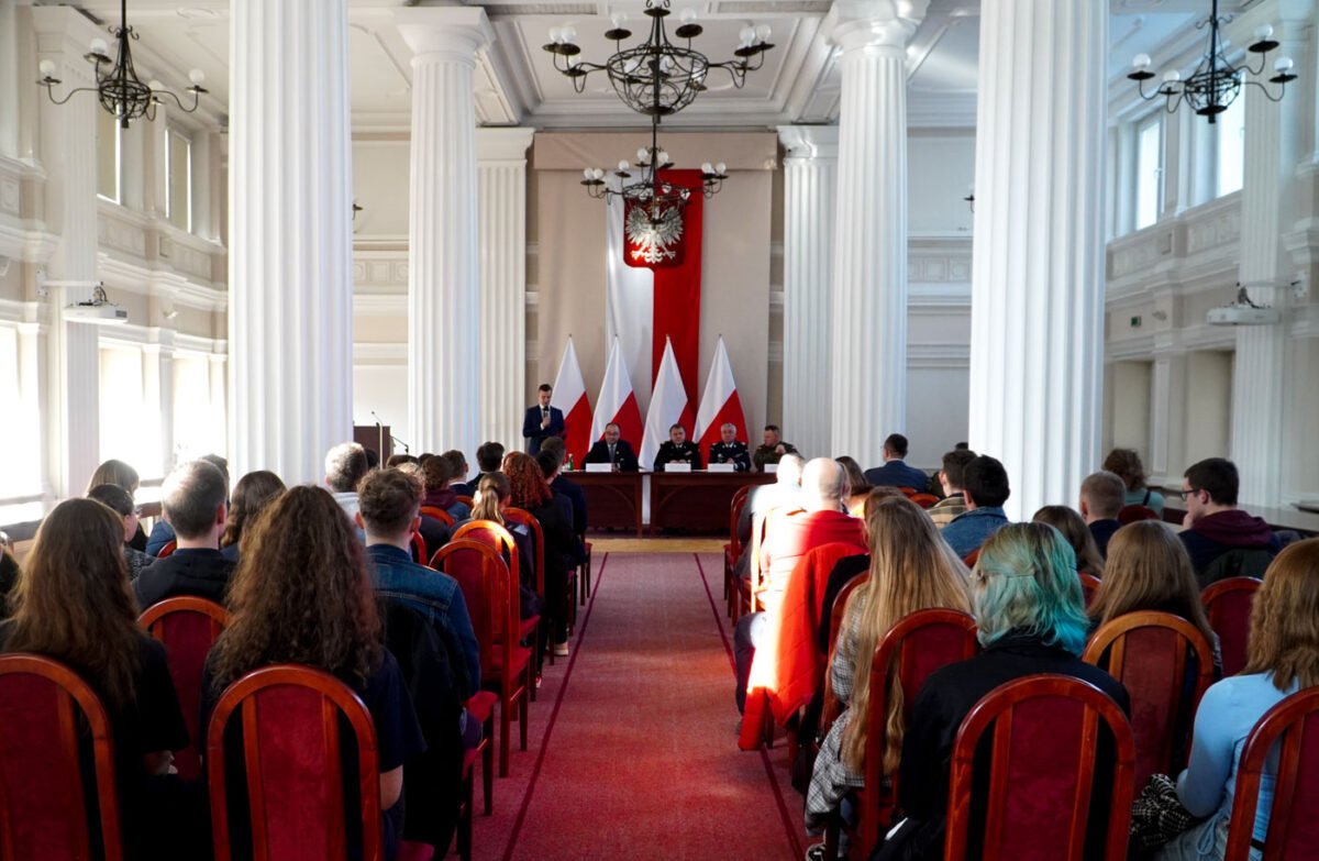 Na zdjęciu widoczna sala konferencyjna z uczestnikami. W dalszej perspektywie zdjęcia widoczny stół prezydialny z nim siedzących czterech mężczyzn i jeden mężczyzna stojący. Trzech mężczyzn ubranych w mundury. Z tyłu za mężczyznami trzy flagi polskie, oraz godło Polski na tle flagi. Przodem do mężczyzn, tyłem do zdjęcia widoczni uczestnicy inauguracji. Widoczne wyposażenie sali cztery żyrandole, dywany oraz kolumny