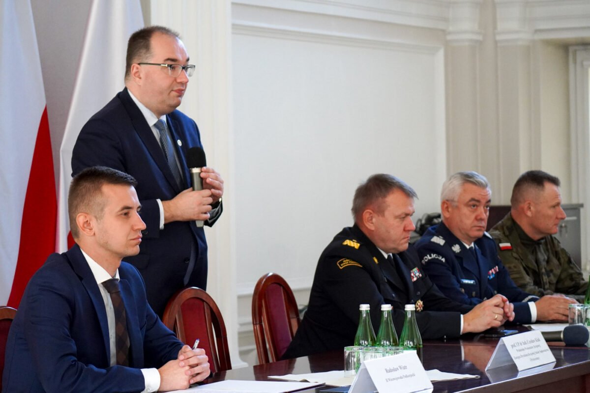 Na zdjęciu widoczna sala konferencyjna. Na pierwszym planie stół prezydialny z nim siedzących czterech mężczyzn i jeden mężczyzna stojący. Trzech mężczyzn ubranych w mundury, dwóch w garniturach. Mężczyzna stojący trzyma w ręku mikrofon