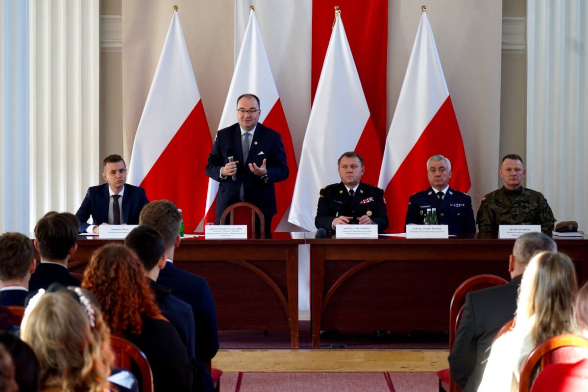 Na zdjęciu widoczna sala konferencyjna z uczestnikami. Na pierwszym planie stół prezydialny z nim siedzących czterech mężczyzn i jeden mężczyzna stojący. Trzech mężczyzn ubranych w mundury. Z tyłu za mężczyznami trzy flagi polskie. Przodem do mężczyzn, tyłem do zdjęcia widoczni uczestnicy inauguracji