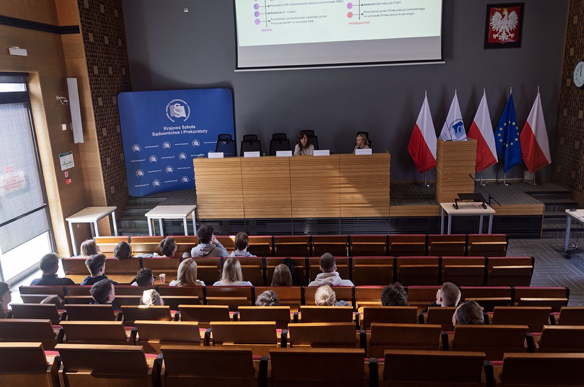 Sala wykładowa. Studenci siedzą na drewnianych krzesłach, przodem do podium, na którym siedzą dwie kobiety. Za nimi stoi flaga Polski i flaga Unii Europejskiej. Po lewej stronie widać banner z logiem Krajowej Szkoły Sądownictwa i Prokuratury. Na ścianie wisi godło Polski, a nad podium jest rzutnik z wyświetlaną prezentacją.