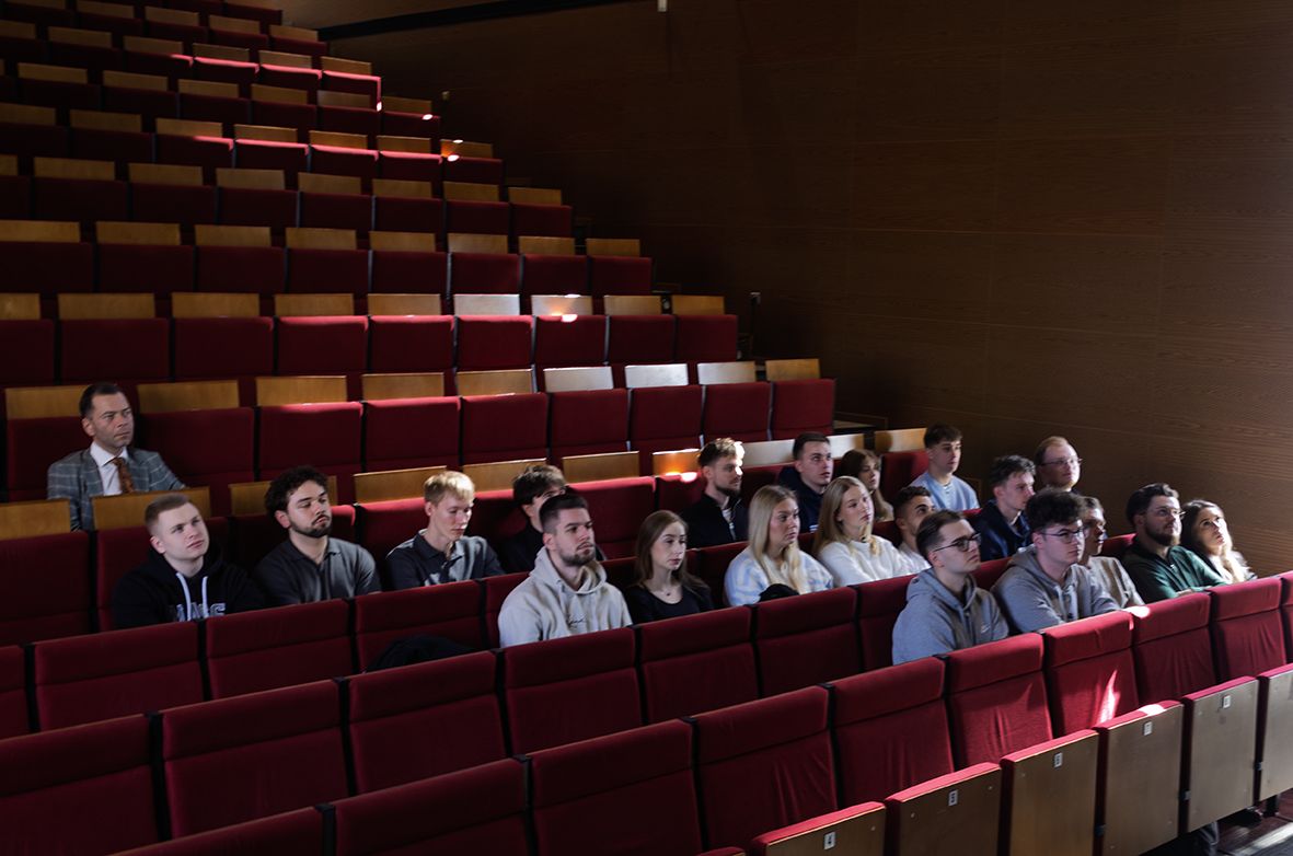 Aula z rzędami czerwonych foteli, na których siedzi grupa ludzi. Osoby te wydają się być w różnym wieku i są różnie ubrane. Aula ma drewniane panele na ścianach. Światło wpada z prawej strony, oświetlając część foteli.