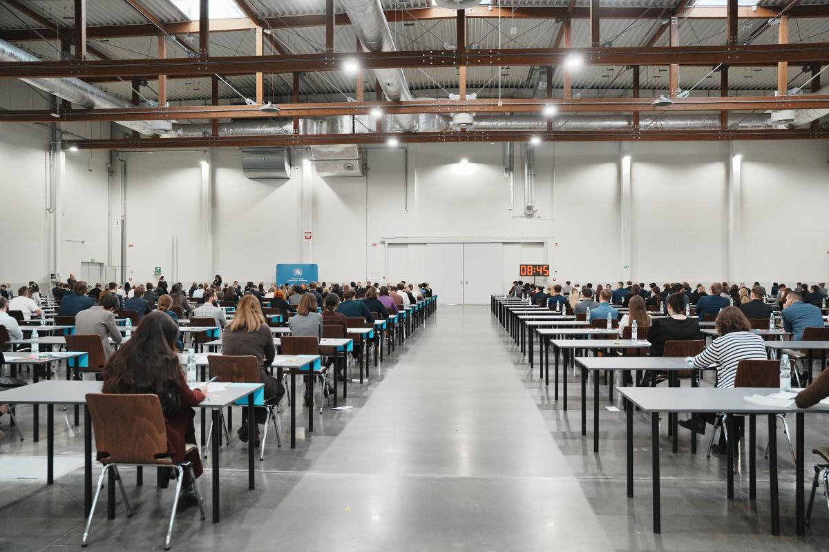 na zdjęciu widać salę, na krzesłach siedzą mężczyźni i kobiety, którzy są uczestnikami konkursu naboru na aplikację. Z przodu sali widać duży zegar elektroniczny oraz baner w kolorze biało-niebieskim z napisem Krajowa Szkoła Sądownictwa i Prokuratury