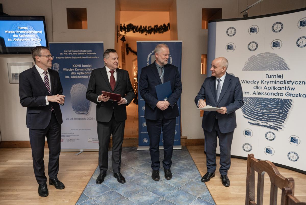 przedstawia wizerunki czterech mężczyzn na tle rollup’ów z logo Instytutu Ekspertyz Sądowych im. dra Jana Sehna oraz Krajowej Szkoły Sądownictwa i Prokuratury oraz XXVII Turnieju Wiedzy Kryminalistycznej im. Aleksandra Głazka 