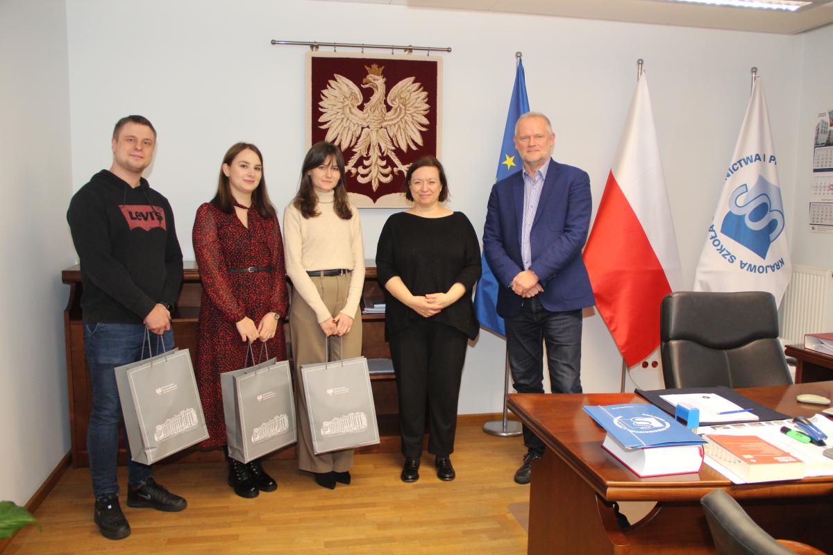 Pięć osób stoi w jasnym pomieszczeniu, po lewej stronie mężczyzna i dwie kobiety trzymają z rękach torby papierowe obok nich kobieta i mężczyzna, wszyscy twarzami zwróconymi w stronę obiektywu. W tle znajdują się trzy flagi, Unii Europejskiej, Polski i Krajowej Szkoły Sądownictwa i Prokuratury. Przed grupą widoczny fragment biurka z fotelem.