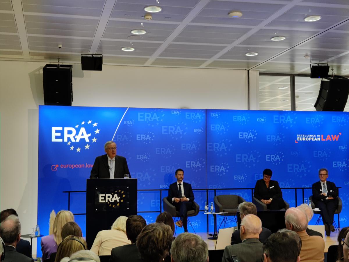 przy mównicy z logo ERA stoi mężczyzna przemawiający do zgromadzonej przed nim publiczności, obok niego na fotelu siedzi mężczyzna, obok jest pusty fotel, obok natomiast siedzi kobieta i kolejny mężczyzna, przed siedzącymi osobami znajduje się stolik z napojami, a za nimi są duże ekrany z niebieskim logo ERA z napisem „Excellence in European Law”