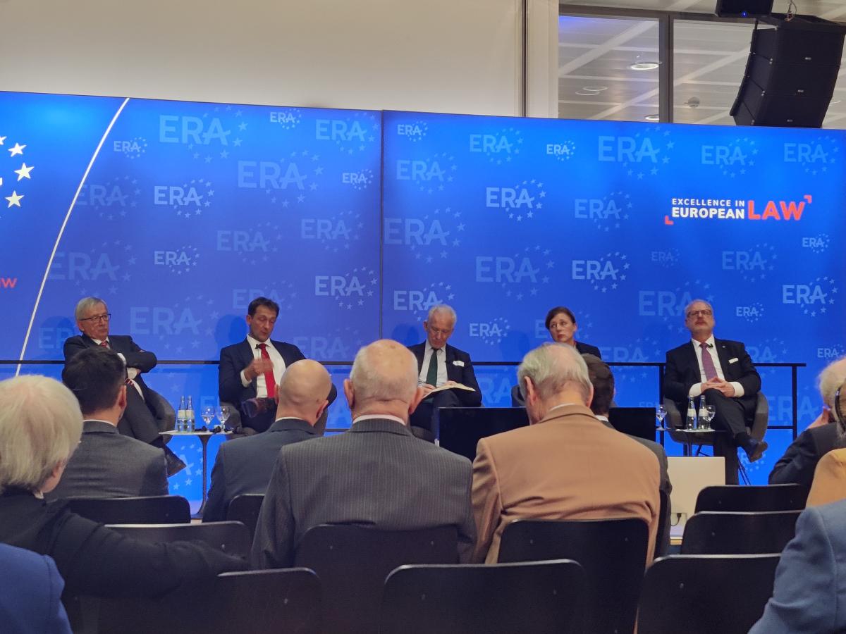 4 mężczyzn i jedna kobieta (druga od prawej) zwróceni przodem do publiczności, siedzący na tle dużych ekranów z niebieskim logo ERA z napisem „Excellence in European Law”, przed nimi stoi stolik z napojami, mężczyźni i kobieta mają przed sobą publiczność siedzącą na krzesłach