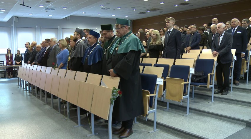 Na zdjęciu widoczna aula z krzesłami. Widoczni stojący kobiety i mężczyźni, z przodu zdjęcia czterech mężczyzn ubranych w togach z insygniami sprawowanych urzędów oraz biretach. Na togach nałożone peleryny w kolorze zielonym, szarym i granatowym oraz na szyi przewieszone