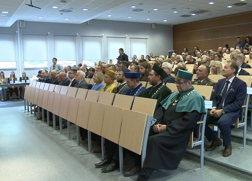 Na zdjęciu widoczna aula z krzesłami. Widoczni siedzący kobiety i mężczyźni, z przodu zdjęcia czterech mężczyzn ubranych w togach z insygniami sprawowanych urzędów oraz biretach. Na togach nałożone peleryny w kolorze zielonym, szarym i granatowym oraz na szyi przewieszone