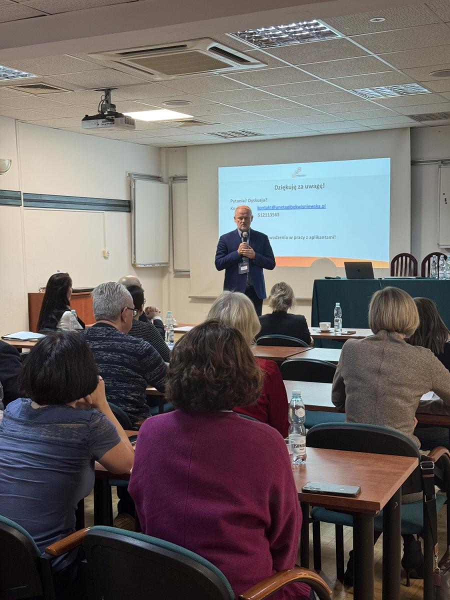 zdjęcie przedstawia stojącego mężczyznę, przemawiającego do siedzących za stołami osób. Mężczyzna trzyma w dłoni mikrofon, za nim widać fragment stołu konferencyjnego, a na ścianie wyświetlana jest pożegnalna prezentacja. Na pierwszym planie widać siedzące za stołami osoby, z twarzami zwróconymi w stronę mówcy. 