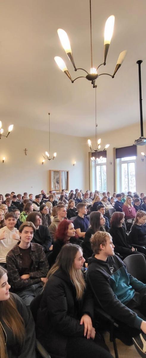 Na zdjęciu widać salę, w której zgromadziła się duża grupa młodzieży, siedzących na krzesłach. Sala jest przestronna, z wysokim sufitem, ozdobiona nowoczesnymi żyrandolami. Na ścianie widoczna jest gablotka. W tle znajdują się duże okna wpuszczające naturalne światło. Atmosfera sugeruje spotkanie lub wykład.