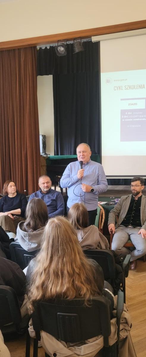 Na zdjęciu widoczna jest scena w sali szkoleniowej lub konferencyjnej. Na pierwszym planie widać osoby siedzące na krzesłach, które uczestniczą w wydarzeniu. Jedna z osób stoi i przemawia, trzymając mikrofon. W tle znajduje się ekran projekcyjny z widocznym napisem „CYKL SZKOLENIA” oraz informacjami dotyczącymi wydarzenia. Po lewej stronie widać zasłonę, a za mówiącą osobą znajduje się pianino lub fortepian. Atmosfera wskazuje na oficjalne spotkanie, takie jak szkolenie lub prelekcja.