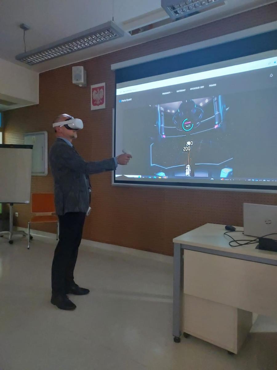Mężczyzna stoi w goglach VR w tle rzutnik z wyświetloną prezentacją.