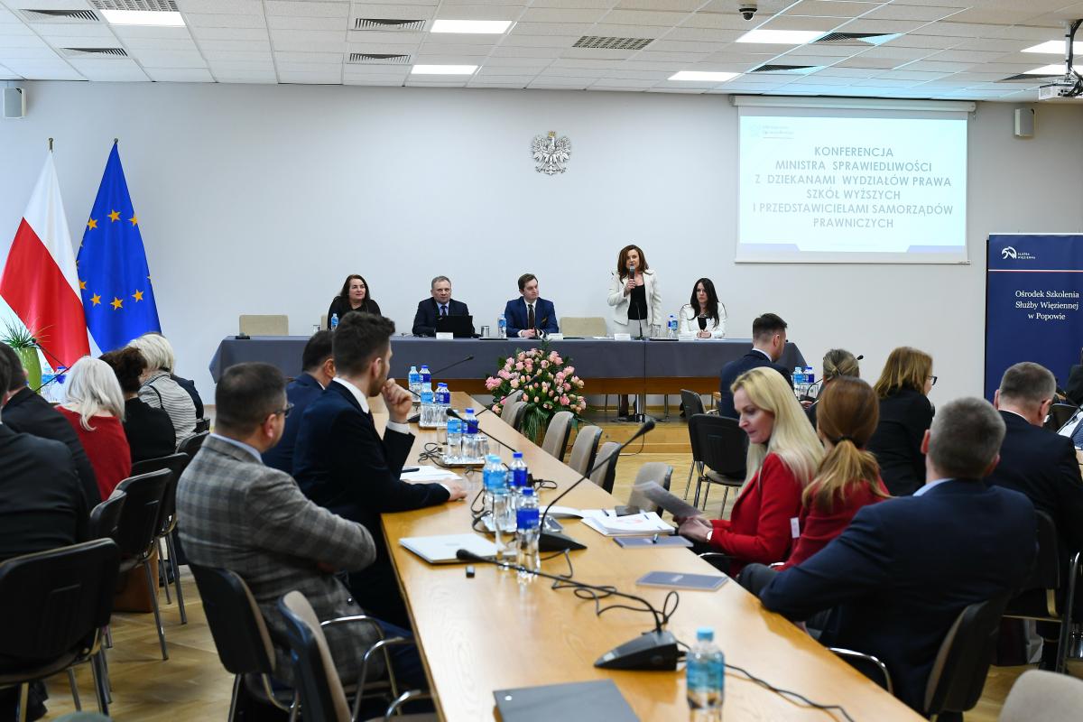 Uczestnicy konferencji, dwóch mężczyzn i trzy kobiety podczas prezentacji przy stole prezydialnym na tle ściany z godłem państwowym, jedna kobieta stoi podczas prezentacji.