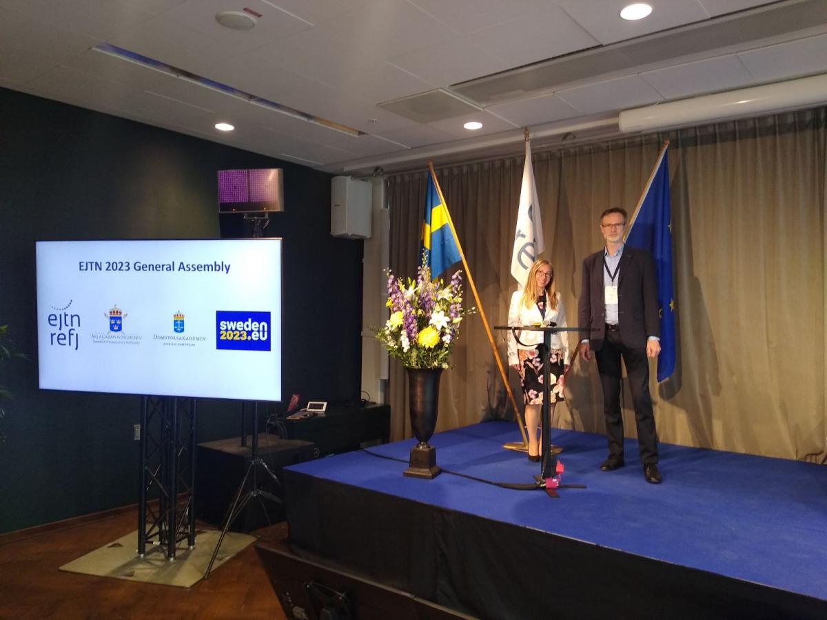 Zdjęcie 1 – przedstawiciele KSSIP pozują do zdjęcia, stojąc na tle obok flag Szwecji, EJTN oraz Unii Europejskiej. Obok na dużym monitorze wyświetlany jest napis z nazwą wydarzenia EJTN 2023 General Assembly oraz logo EJTN i szwedzkiej prezydencji w UE. 