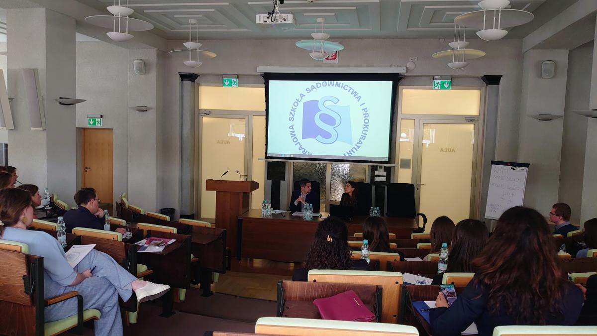 Sala wykładowa podczas szkolenia na której siedzą uczestnicy odwróceni w stronę prezentacji wyświetlanej na białym rzutniku, na którym widnieje logo KSSiP. Kobieta i mężczyzna siedzą przy stole prezydialnym