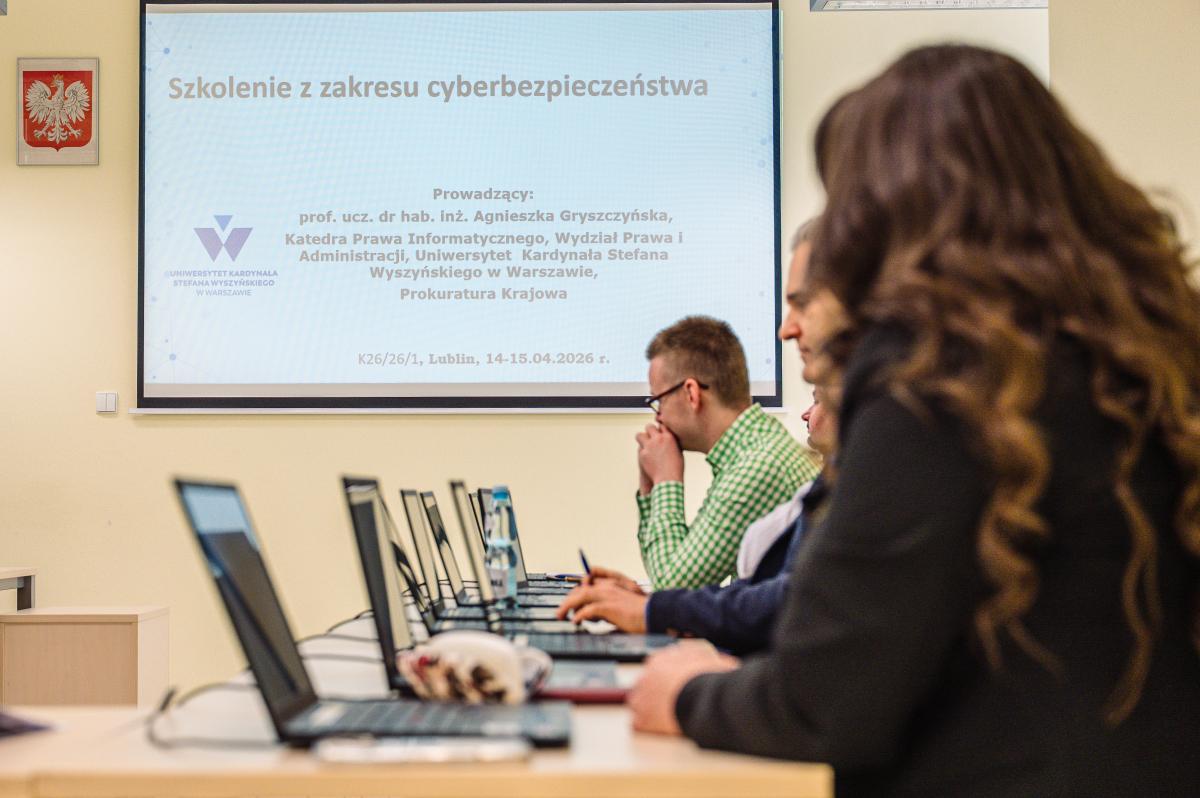 Zdjęcie zrobione podczas zajęć warsztatowych szkolenia z cyberprzestępczości. Na pierwszym planie uczestnic siedzący bokiem, prostopadle do tablicy, na której wyświetlana jest prezentacja (na prezentacji – tytuł szkolenia i dane prowadzącego). Przed każdym z uczestników stoi otworzony laptop.