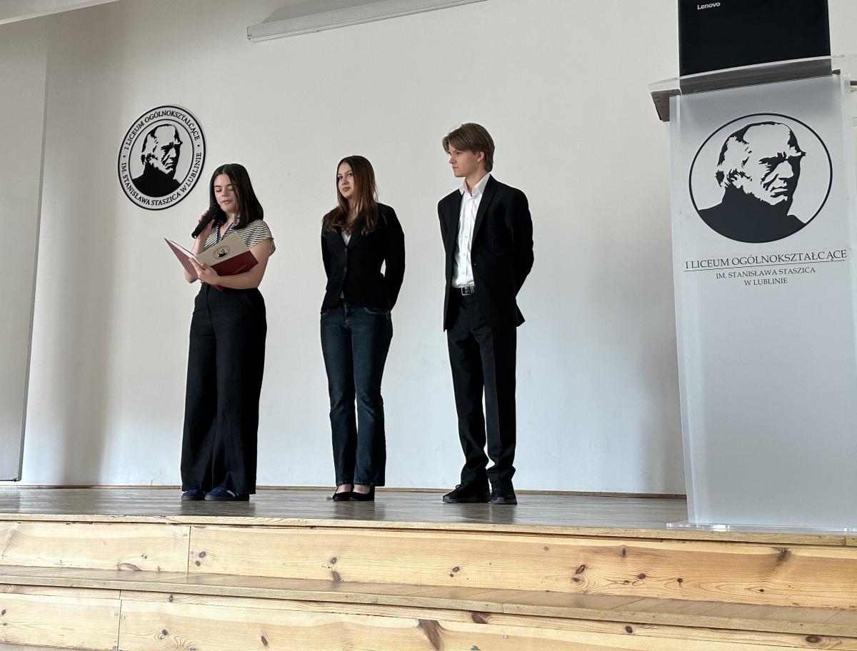 Zdjęcie przedstawia trzy młode osoby – licealistów, dwie dziewczyny i chłopak. Stoją na środku sceny. Za nimi jest biała ściana z logo. Logo zawiera obraz twarzy Stanisława Staszica oraz tekst: "Pierwsze Liceum Ogólnokształcące imienia Stanisława Staszica w Lublinie." Po prawej stronie zdjęcia znajduje się mównica. Na jej froncie znajduje się takie samo logo.