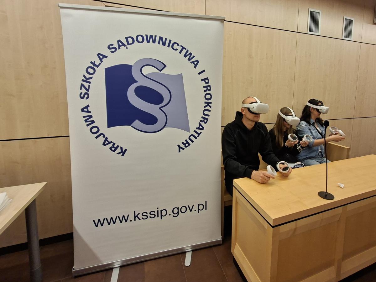 Warsztaty VR podczas wydarzenia Noc Otwartych Sądów. Trzy osoby siedzą przy stole w sali rozpraw, mają na głowach gogle VR i trzymają kontrolery. Obok nich stoi roll-up z logo Krajowej Szkoły Sądownictwa i Prokuratury oraz adresem internetowym www.kssip.gov.pl.