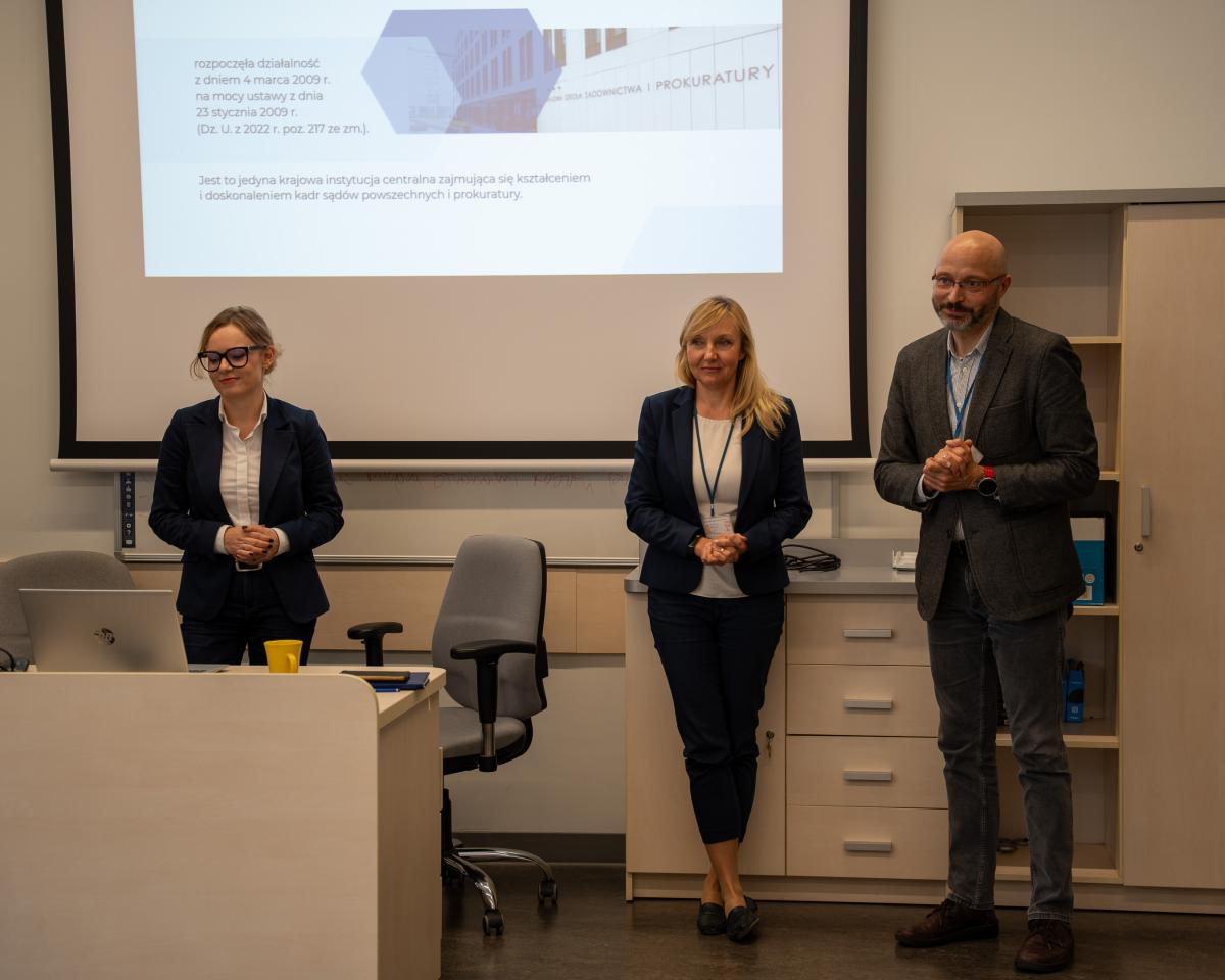 trzy osoby: jeden mężczyzna i dwie kobiety stoją przed tablicą z wyświetloną prezentacją dotyczącą Krajowej Szkoły Sądownictwa i Prokuratury.Przed jedną z kobiet stoi biurko. Na biurku znajduje się żółty kubek.