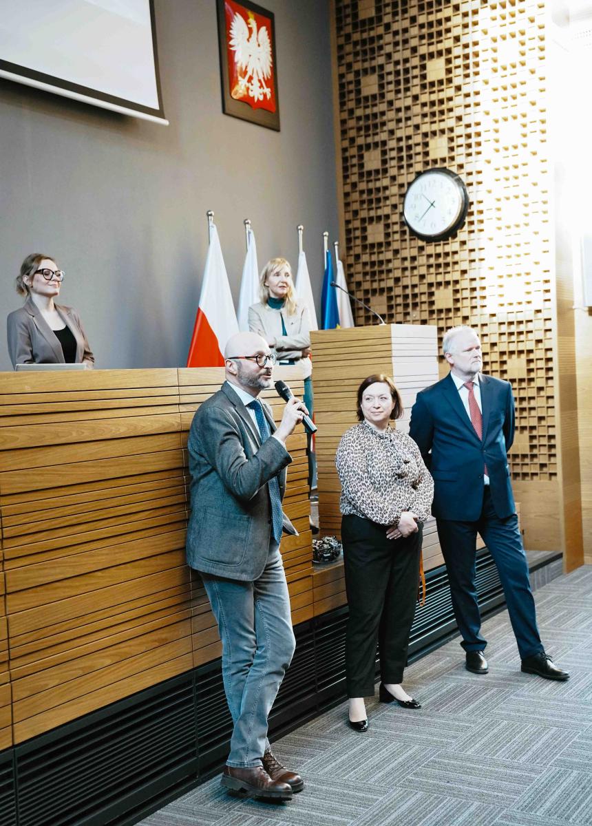 dwie kobiety stoją na podwyższeniu, przy dużym pulpicie. Poniżej stoją dwaj mężczyźni i kobieta. Jeden z mężczyzn trzyma w ręku mikrofon. W tle na ścianie widać godło Polski i zegar. 