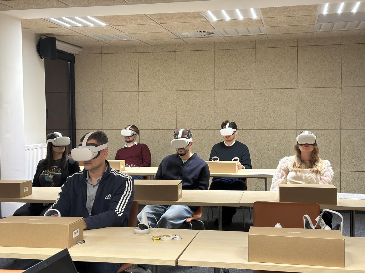 Na zdjęciu widoczna jest sala szkoleniowa z uczestnikami siedzącymi przy stołach. Na stołach leżą kartonowe pudełka oraz zestawy urządzeń do wirtualnej rzeczywistości. Osoby korzystają z gogli VR. Pomieszczenie ma jasne oświetlenie i neutralny wystrój.