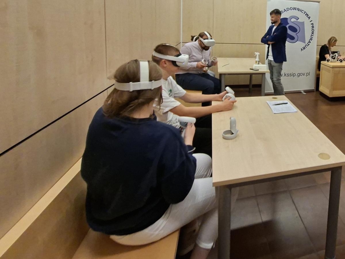 Warsztaty VR podczas wydarzenia Noc Otwartych Sądów. Cztery osoby siedzące przy stołach w sali rozpraw uczestniczą w pokazie VR – mają założone gogle VR i trzymają kontrolery. W tle stoi prowadzący warsztaty ubrany w granatową marynarkę i szare jeansy oraz roll-up z logo Krajowej Szkoły Sądownictwa i Prokuratury.