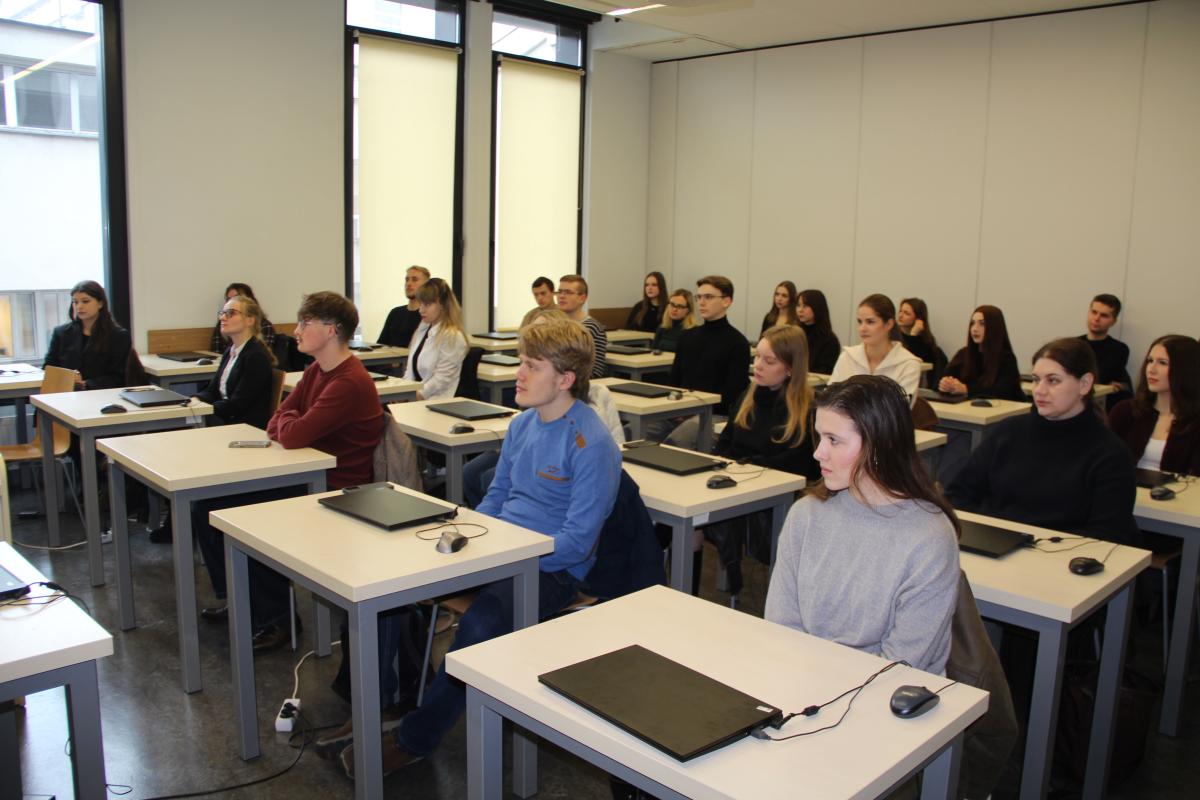 Grupa mężczyzn i kobiet siedzi przy stolikach. Na większości stolików leżą laptopy. Naprzeciwko grupy kobiet i mężczyzn ustawione jest biurko z laptopem, przy którym siedzą 2 kobiety. W tle widoczne duże okna, za którymi znajduje się kolejny budynek.