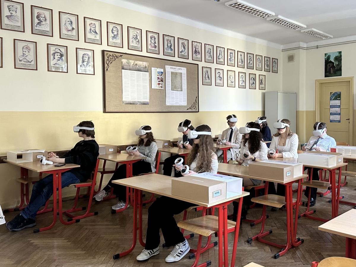 Zdjęcie przedstawia salę lekcyjną z kilkunastoma uczniami siedzącymi przy pojedynczych ławkach. Wszyscy uczniowie mają na głowach gogle VR (wirtualnej rzeczywistości) i trzymają w rękach kontrolery. Uczestniczą w zajęciach edukacyjnych z wykorzystaniem technologii VR. Sala ma klasyczny wystrój – na ścianach wiszą portrety historycznych postaci, a podłoga jest wyłożona drewnianym parkietem. Uczniowie są skupieni na doświadczeniu wirtualnym, a ich postawa świadczy o zaangażowaniu.