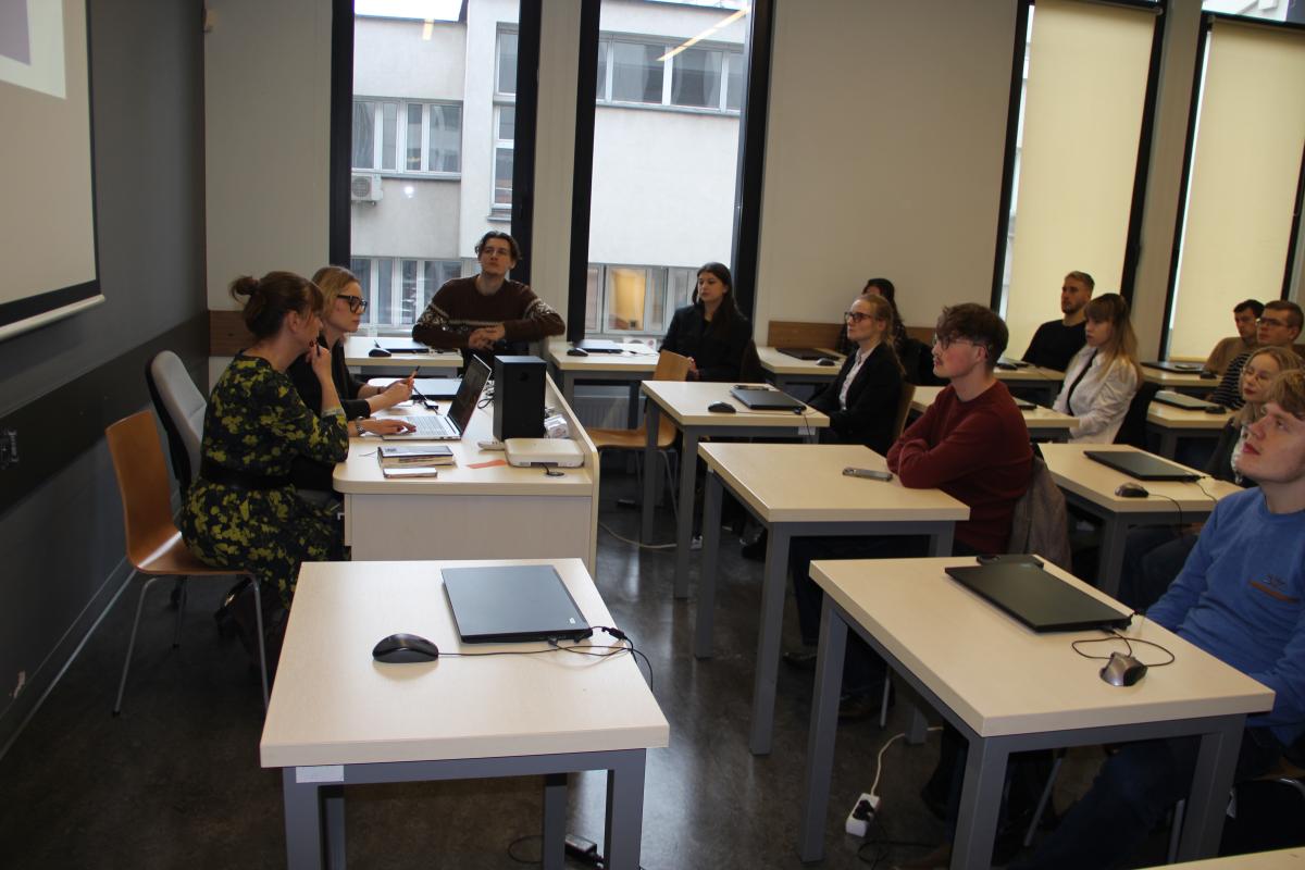 Grupa mężczyzn i kobiet siedzi przy stolikach. Na większości stolików leżą laptopy. W tle widoczne trzy duże okna. Dwa okna są zasłonięte roletami.