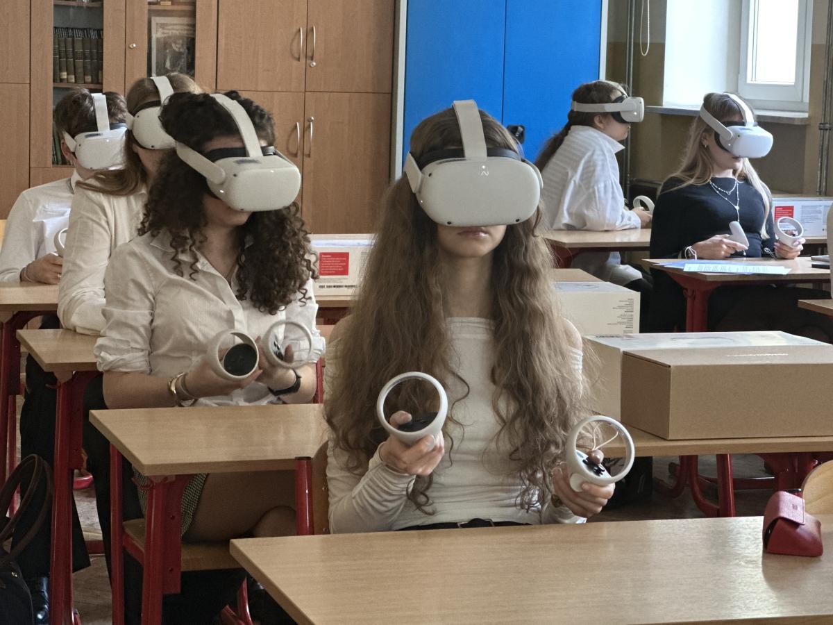 Zdjęcie przedstawia klasę, w której uczniowie siedzą przy drewnianych biurkach. Mają na głowach zestawy do wirtualnej rzeczywistości (VR), trzymają kontrolery VR. Uczniowie wydają się być zaangażowani w aktywność z użyciem sprzętu VR. Klasa ma drewniane szafki, niebieskie drzwi szafki oraz okna, przez które wpada naturalne światło.