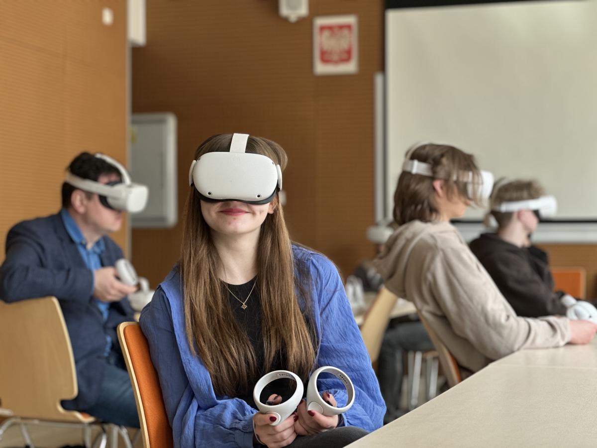 Zdjęcie przedstawia cztery osoby siedzące w sali wykładowej. Każda osoba ma na sobie zestaw do wirtualnej rzeczywistości, składający się z gogli VR na głowach i kontrolerów VR w rękach. W tle widać białą tablicę oraz kilka krzeseł. Pomieszczenie jest jasne i dobrze oświetlone.