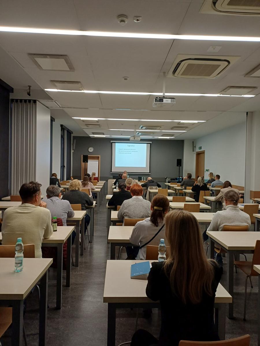 Na zdjęciu uczestnicy szkolenia podczas zajęć. Zdjęcie zrobione  na tyłach w sali wykładowej, widać tylko plecy uczestników. W głębi  sali widać tablice z wyświetlana prezentacją.