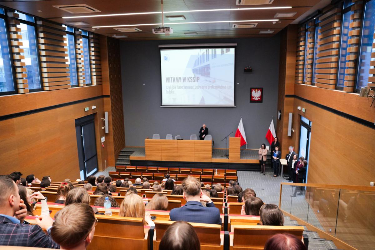 Zdjęcie przedstawia powitanie aplikantów XVII rocznika aplikacji prokuratorskiej odbywające w auli KSSiP. Uczestnicy siedzą na widowni, twarzami zwróceni w stronę sceny. Na scenie na podwyższeniu przy mównicy stoi mężczyzna, za którym znajduje się ekran powitalny oraz dwie flagi Polski. Na ekranie widoczny jest slajd z napisem „Witamy w KSSiP, czyli jak funkcjonować , żeby było profesjonalnie i po ludzku”. Po lewej stronie mężczyzny, przy podwyższeniu, stoi grupa siedmiu osób, twarzami zwrócona w stronę audytorium. 