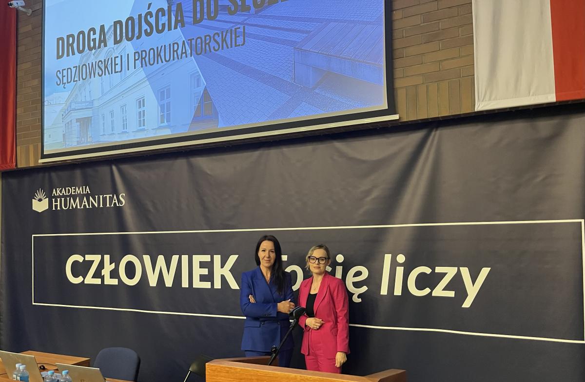 widok sali z dużym rzutnikiem, na  którym wyświetlony jest napis: Droga dojścia do służby sędziowskiej i prokuratorskiej. Poniżej znajduje się napis Akademia Humanitas. Człowiek. To się liczy. Z przodu stoi długi drewniany stół oraz miejsce do przemawiania, na których umieszczone jest godło Polski w złotym kolorze. Za stołem stoją dwie kobiety.