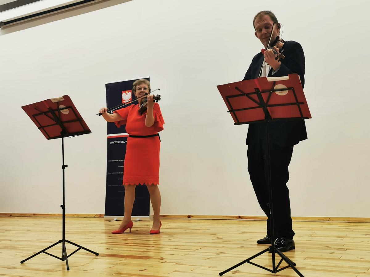 Na zdjęciu widać dwie osoby stojące na drewnianej scenie w jasnym pomieszczeniu. Oboje grają na skrzypcach i stoją obok siebie, zwróceni w stronę widza. Po lewej stronie znajduje się kobieta ubrana w elegancką, czerwoną sukienkę i czerwone buty na obcasie. Po prawej stronie stoi mężczyzna w ciemnym garniturze, białej koszuli i czarnych butach. Przed każdą z osób ustawiony jest pulpit z nutami. W tle, za kobietą, widoczny jest stojący baner z godłem Polski. Całość odbywa się na jasnej, drewnianej podłodze, a ściana za muzykami jest gładka i jasna.