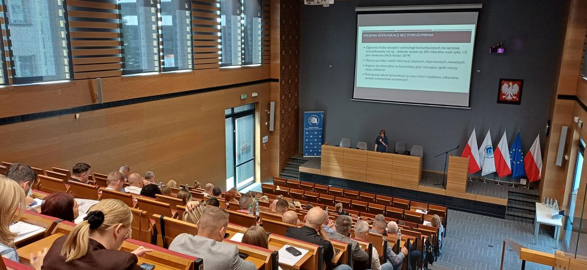 Grupa około sześćdziesięciu osób: kobiet i mężczyzn siedzi na dużej auli wykładowej. Przed nimi, na podeście, przy dużym biurku stoi kobieta. Nad głową kobiety znajduje się duża tablica z wyświetloną prezentacją. Tytuł wyświetlanego slajdu to: Epidemia komunikacji bez porozumienia. W tle po prawej stronie widać godło Polski oraz flagi Polski, Unii Europejskiej i Krajowej Szkoły Sądownictwa i Prokuratury. Po lewej stronie znajduje się roll-up z logo Krajowej Szkoły Sądownictwa i Prokuratury.