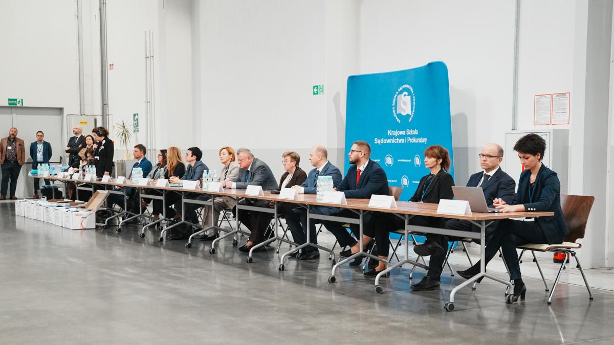 kobieta z mikrofonem w ręce przemawia do uczestników konkursu, obok siedzi dziewięć kobiet i pięciu mężczyzn za stołem. Przed sobą mają na stole ustawione tabliczki z imieniem i nazwiskiem. Za ich plecami ustawiony jest baner w kolorze biało-niebieskim z napisem Krajowa Szkoła Sądownictwa i Prokuratury. Obok stołu po lewej stronie stoi jedna kobieta i trzech mężczyzn.