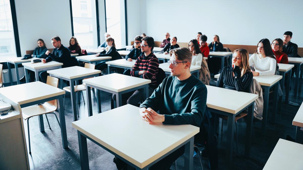 grupa dziewiętnastu mężczyzn i kobiet siedzi w sali przy stolikach. Są oni zwróceni twarzami w stronę obiektywu. Po ich prawej stronie znajdują się duże okna. 