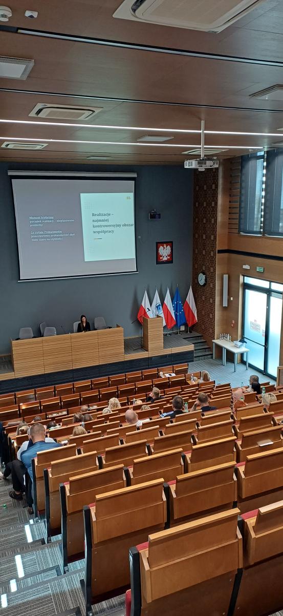 Grupa około sześćdziesięciu osób: kobiet i mężczyzn siedzi na dużej auli wykładowej. Przed nimi, na podeście, przy dużym biurku siedzi kobieta. Nad głową kobiety znajduje się duża tablica z wyświetloną prezentacją. Tytuł wyświetlanego slajdu to: Realizacje - najmniej kontrowersyjny obszar współpracy. W tle po prawej stronie widać godło Polski oraz flagi Polski, Unii Europejskiej i Krajowej Szkoły Sądownictwa i Prokuratury. Po lewej stronie znajduje się roll-up z logo Krajowej Szkoły Sądownictwa i Prokuratury.