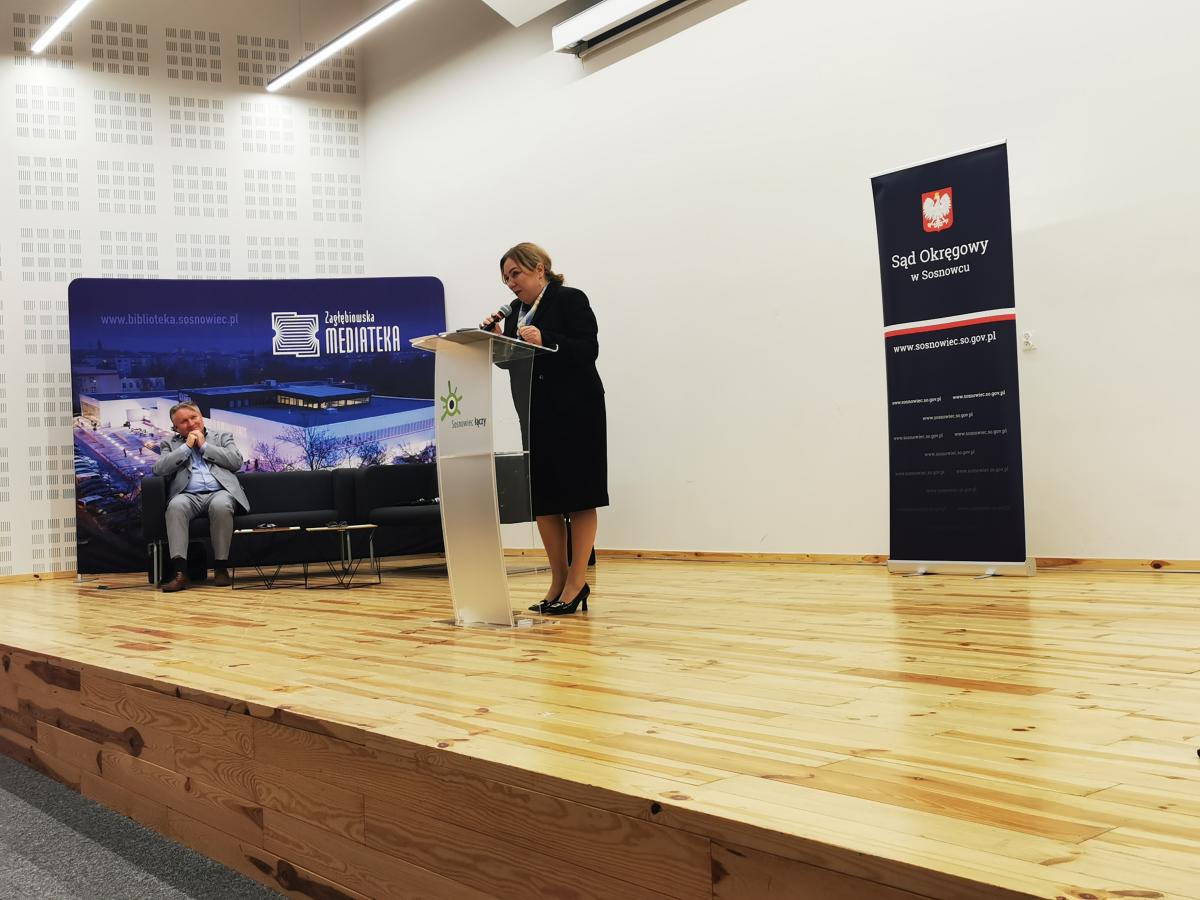 Na zdjęciu widoczna jest kobieta stojąca na drewnianej scenie i przemawiająca do mikrofonu przy przezroczystej mównicy. Ubrana jest w elegancki, czarny strój oraz czarne buty na obcasie. Po lewej stronie sceny, na tle dużego niebieskiego baneru z napisem „Zagłębiowska Mediateka” i zdjęciem budynku, siedzi mężczyzna w jasnym garniturze na ciemnej kanapie. Po prawej stronie sceny ustawiony jest pionowy baner z godłem Polski i napisem „Sąd Okręgowy w Sosnowcu”. Całość odbywa się w jasnym, przestronnym pomieszczeniu.