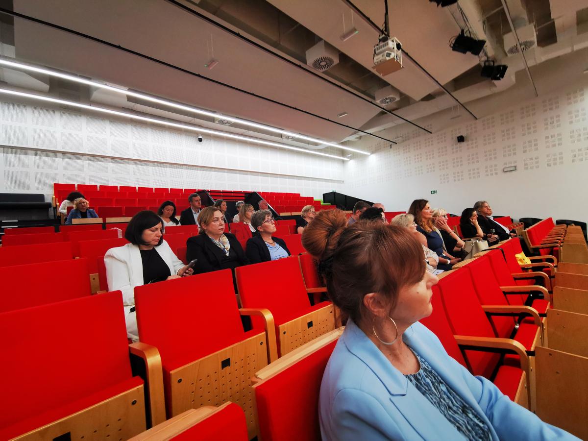 Na zdjęciu widać scenę w nowoczesnej sali konferencyjnej z jasnym, drewnianym podestem. Po lewej stronie sceny stoi czarna kanapa, na której siedzi mężczyzna w garniturze. Obok kanapy znajduje się niski, czarny stolik. Za kanapą ustawiona jest duża plansza z napisem „Zagłębiowska Mediateka” i zdjęciem nowoczesnego budynku biblioteki. Na środku sceny stoi mównica z przezroczystego materiału. Przy mównicy stoi mężczyzna w marynarce, przemawiający do mikrofonu. Po prawej stronie sceny ustawiony jest pionowy baner z napisem „Sąd Okręgowy w Sosnowcu” oraz godłem Polski. Tło sali jest jasne, a na suficie wiszą nowoczesne, skośne lampy. Całość sprawia wrażenie oficjalnego spotkania lub konferencji.